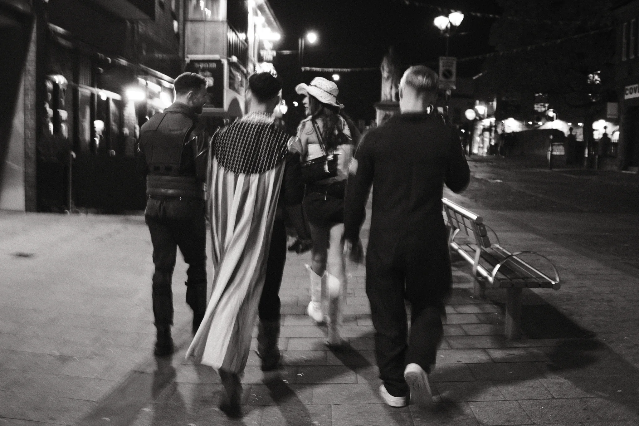A group walks away through Ormskirk, with a pale cape and a light hat catching the street lighting.