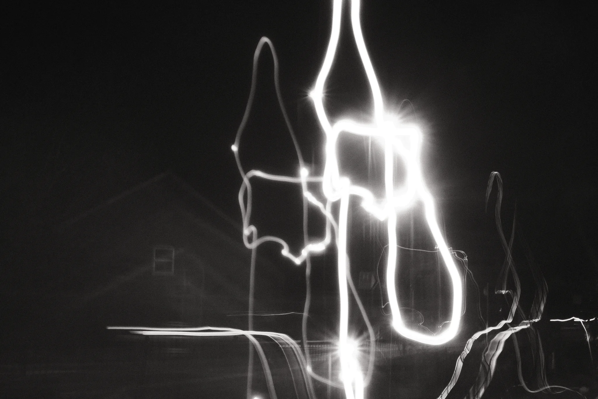 Long white light trails loop across a night scene, with the faint outline of a house behind them.