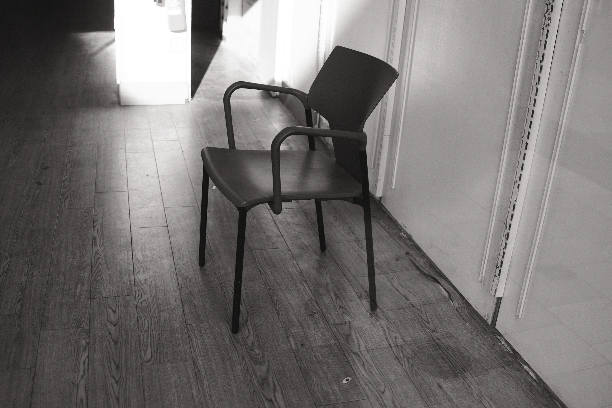 An empty chair sits beside an open doorway, with pale walls and diagonal floorboards around it.