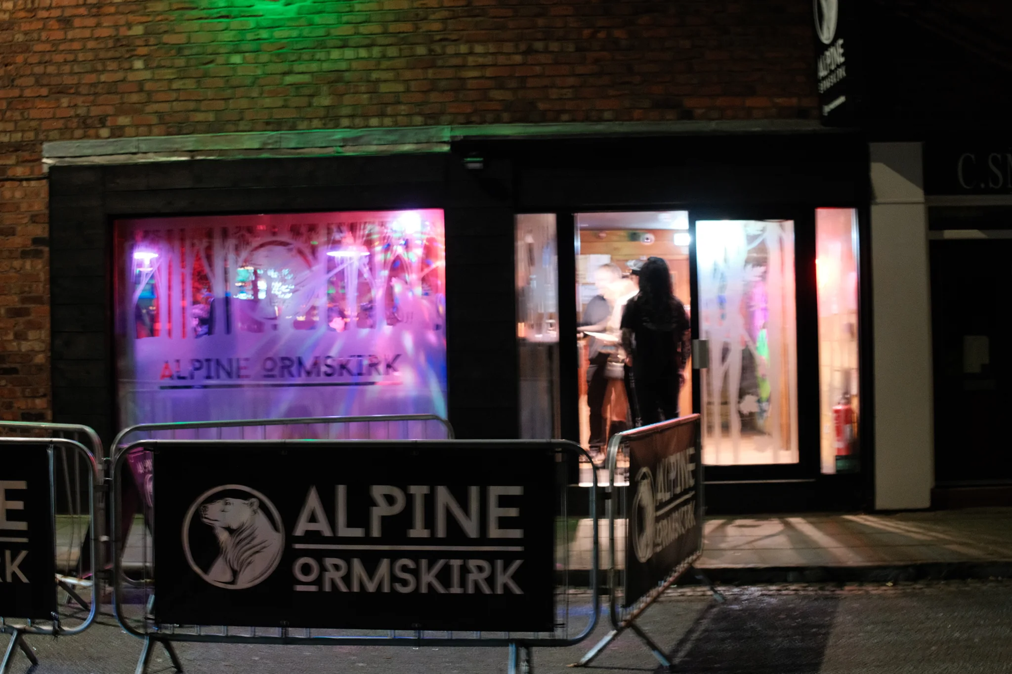 Pink, violet and green light surround a dark figure in the doorway of Alpine Ormskirk.