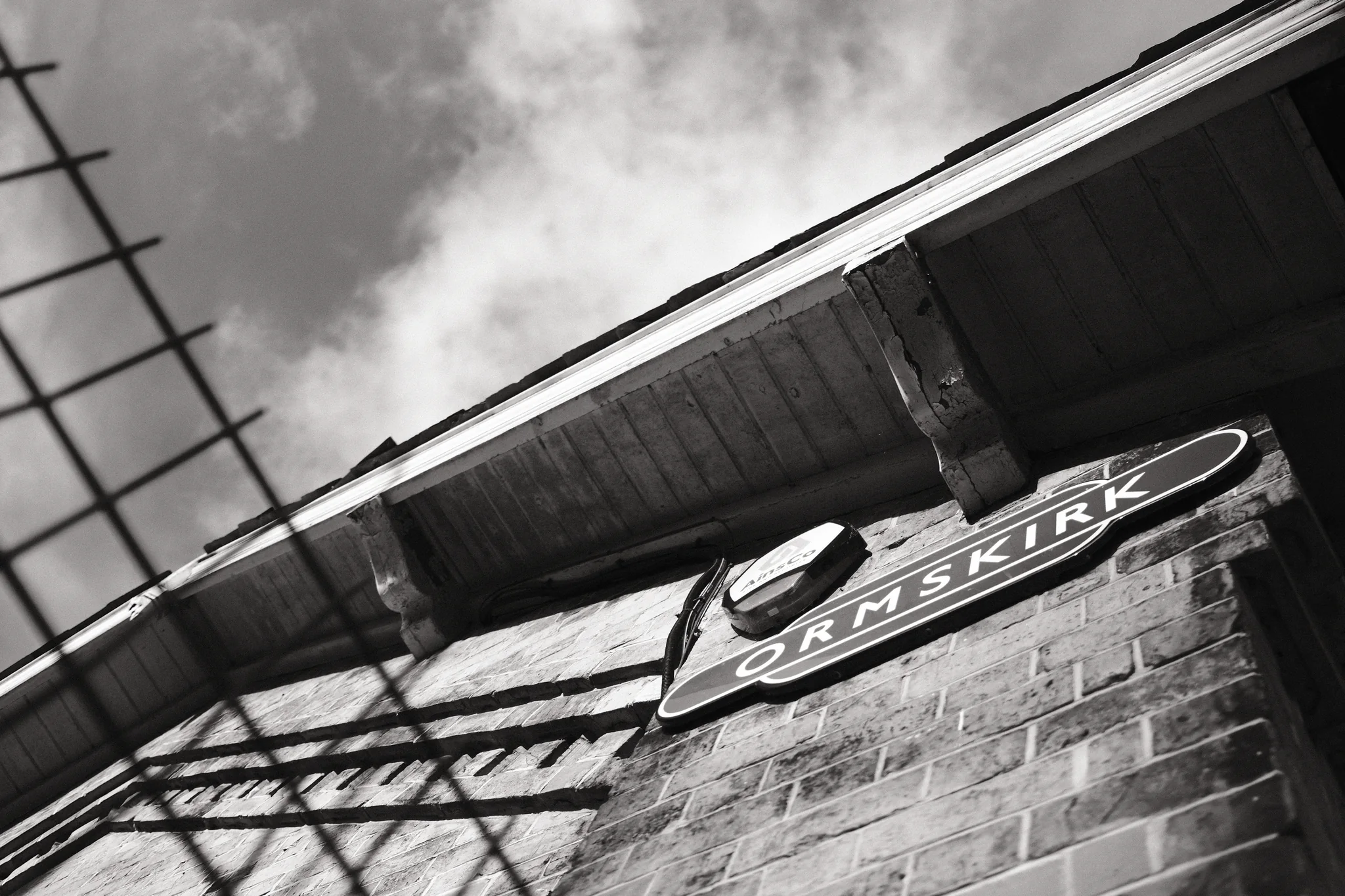 The Ormskirk station sign and roofline run diagonally across the frame, behind a blurred foreground fence.