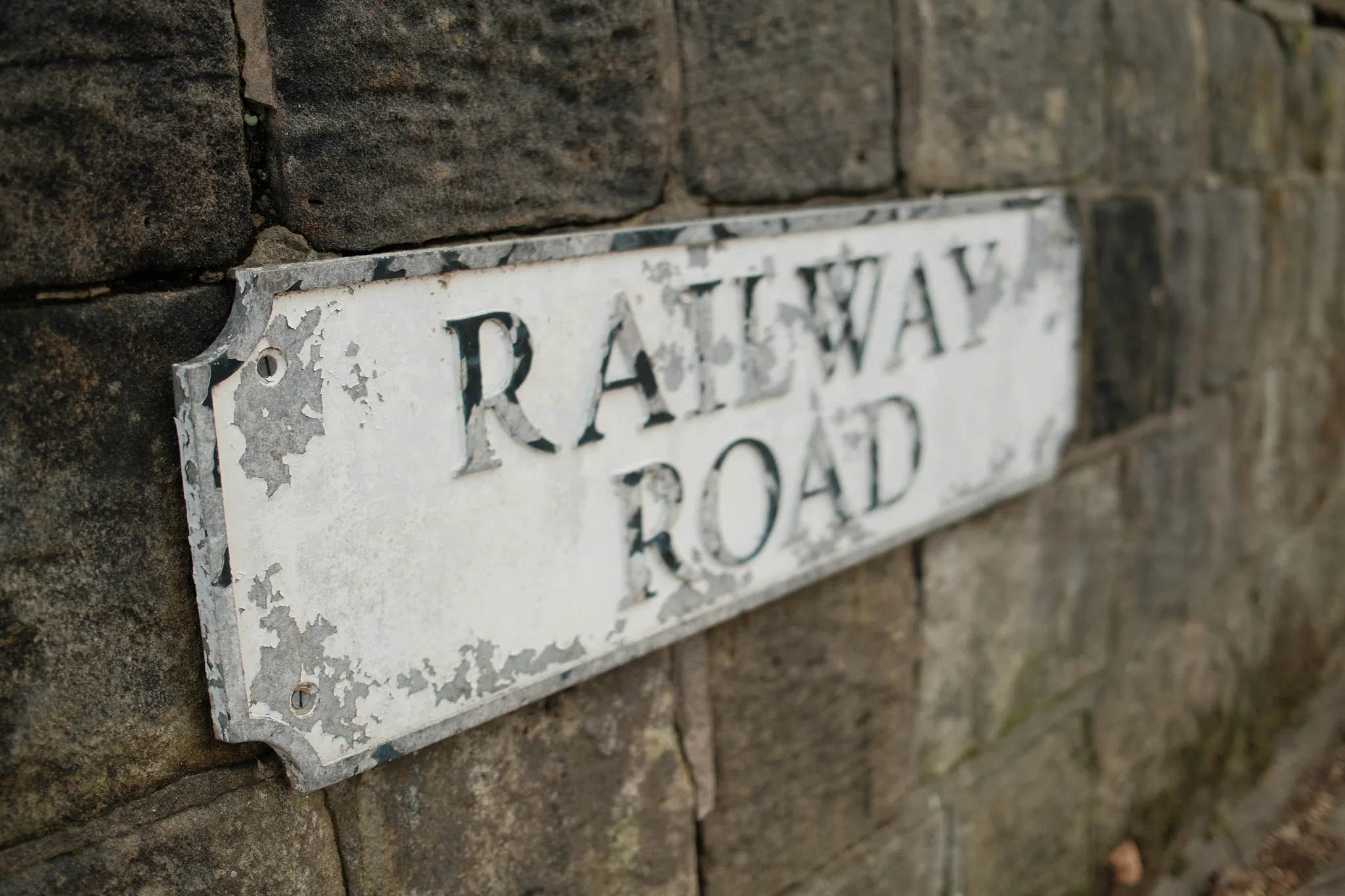 Flaking paint and dark lettering cover a Railway Road sign mounted on a stone wall.