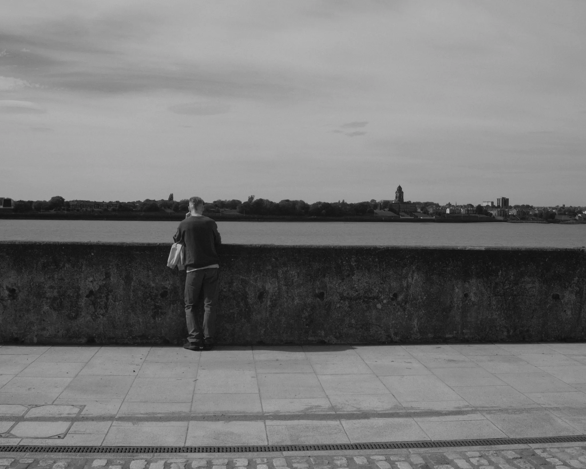 A lone person standing at a waterfront wall, in black and white.