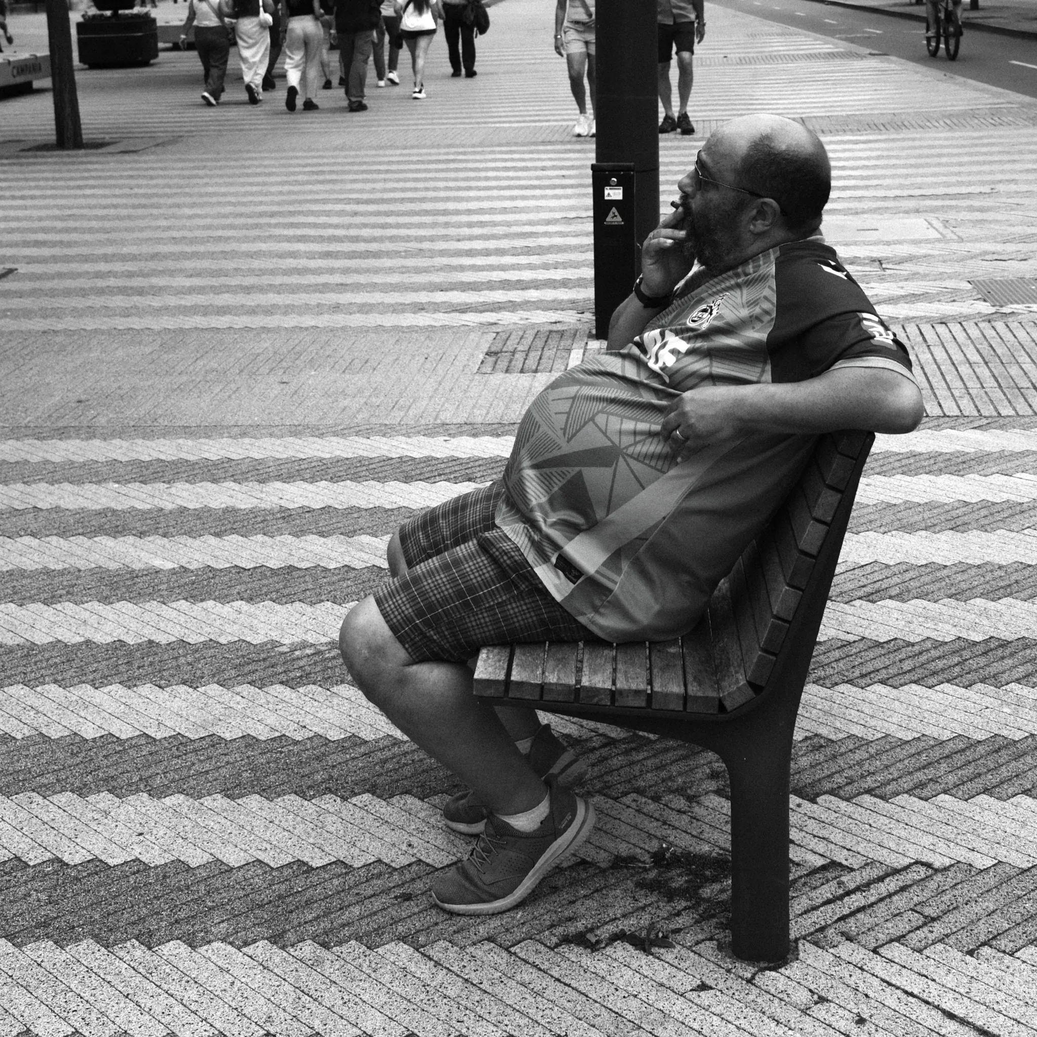 A seated person leaning back on a bench beside a patterned pavement, in black and white.
