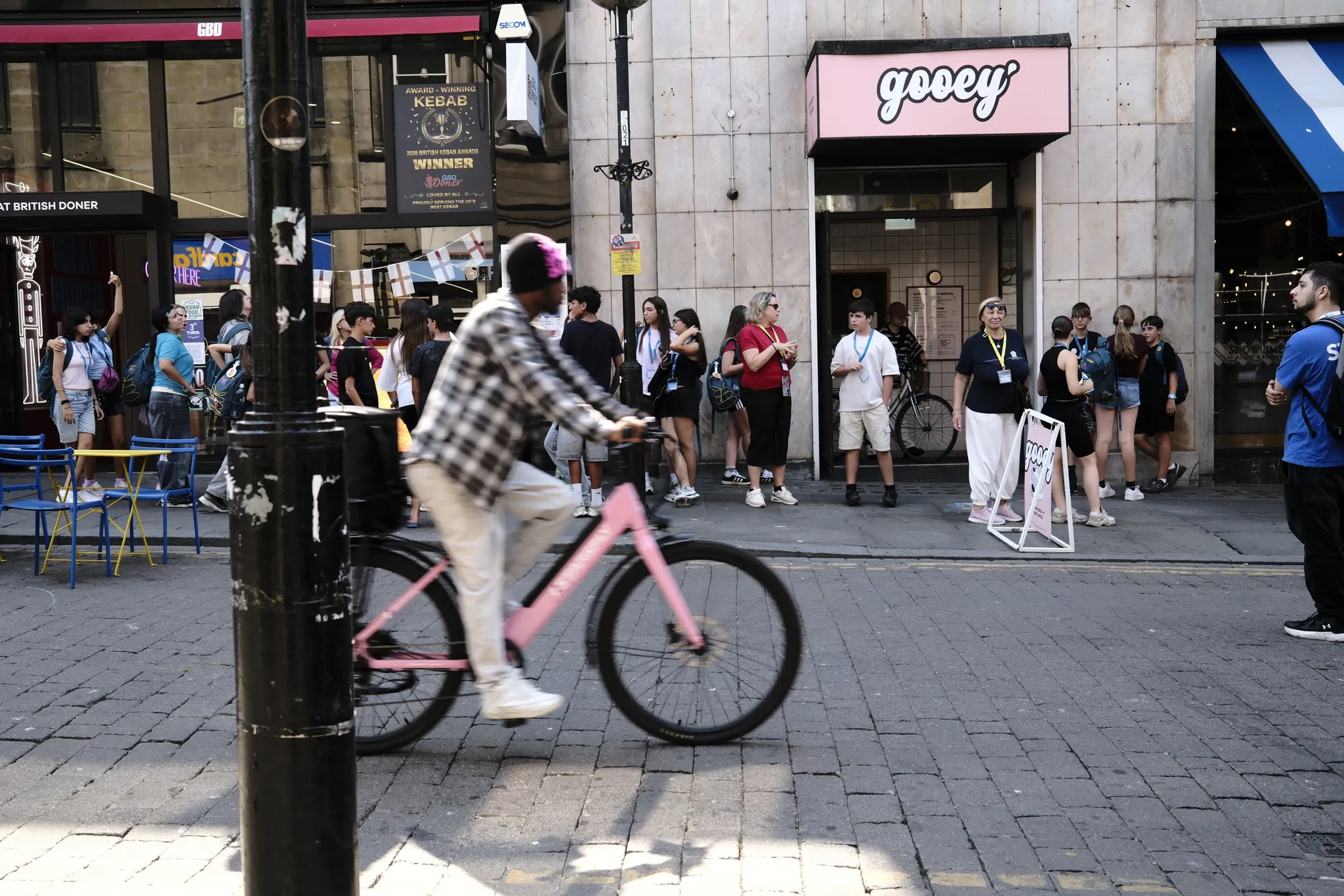 A cyclist on a pink bicycle passes café tables and the Gooey shopfront on Bold Street.