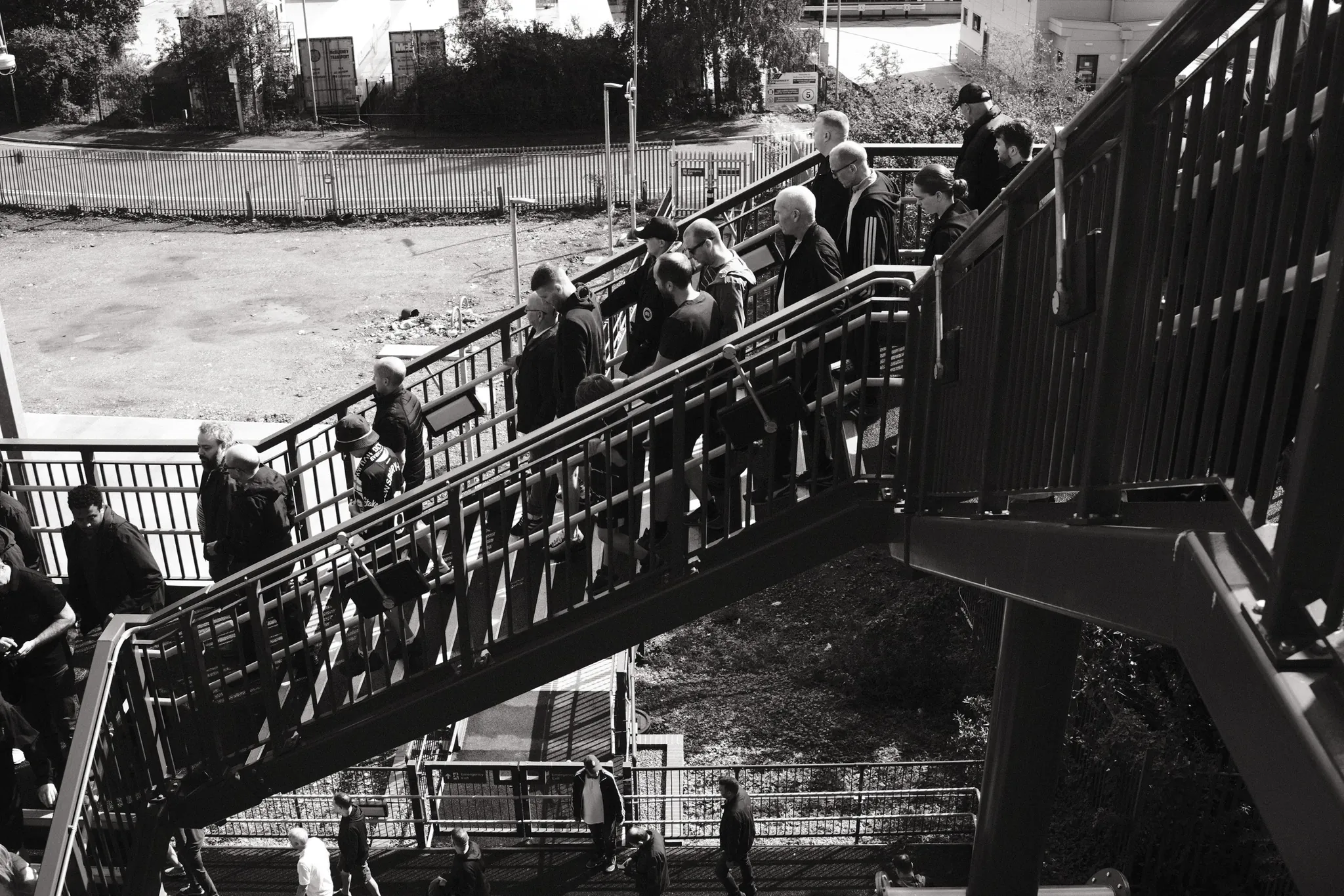 People descend a metal footbridge staircase beside railway infrastructure in black and white.