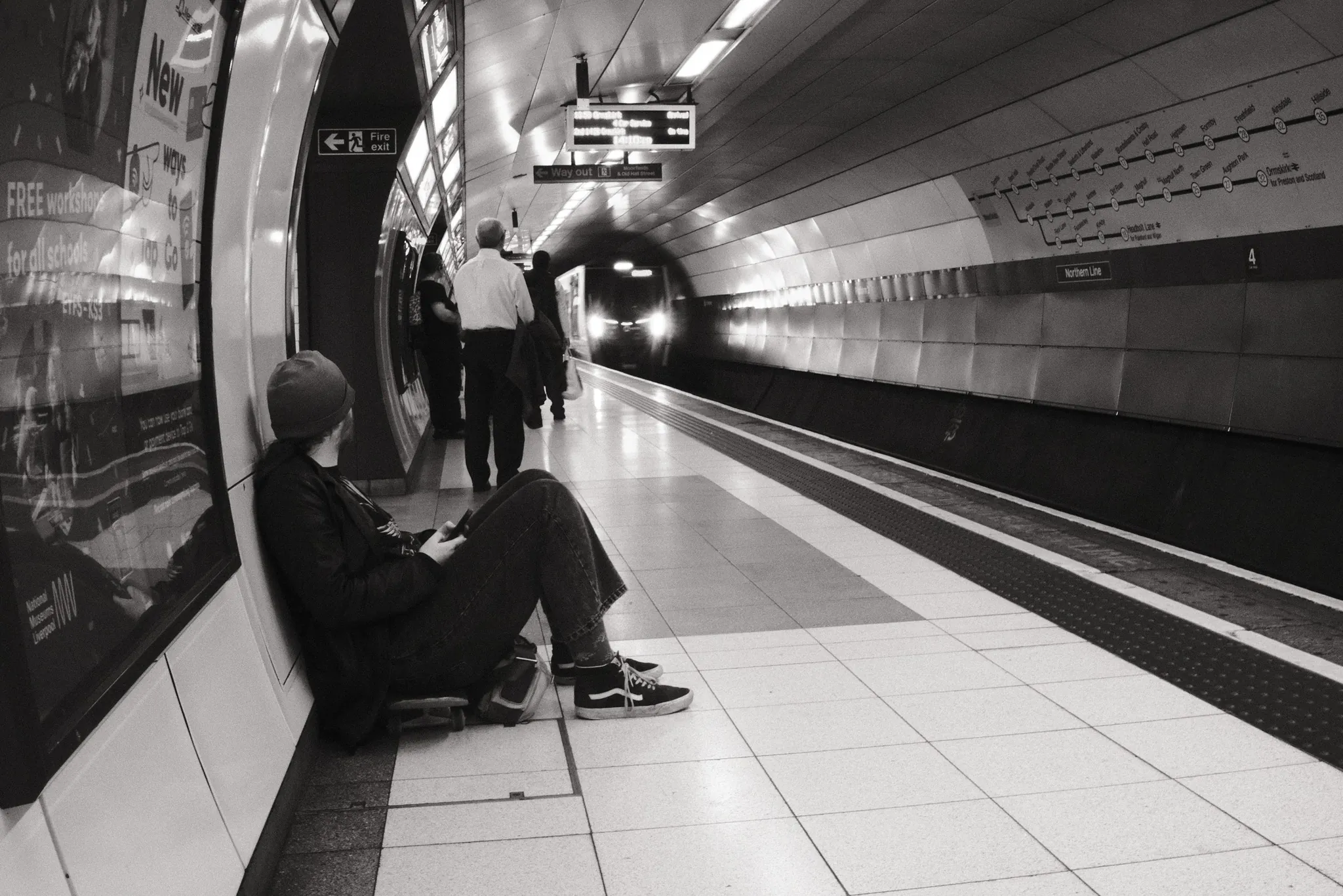A seated passenger waits on a Moorfields platform as a train approaches through the tunnel.