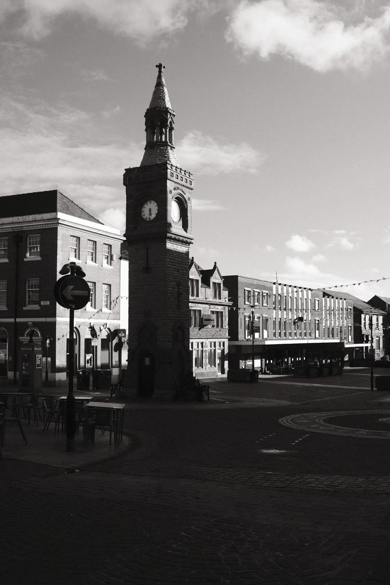 Ormskirk Clock Tower and surrounding shops stand beneath a cloudy sky in black and white.