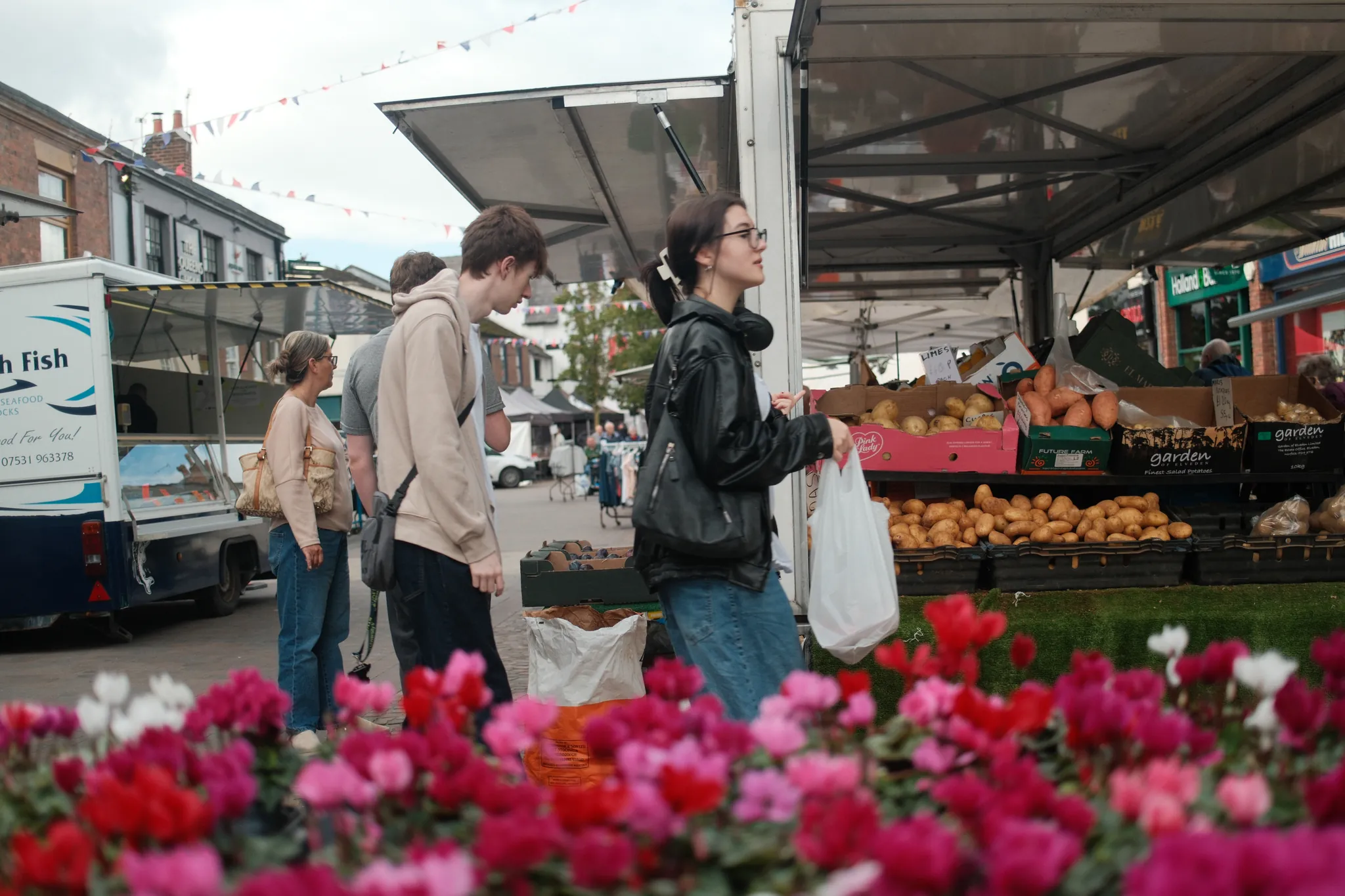Bright flowers fill the foreground as shoppers pass a produce stall.