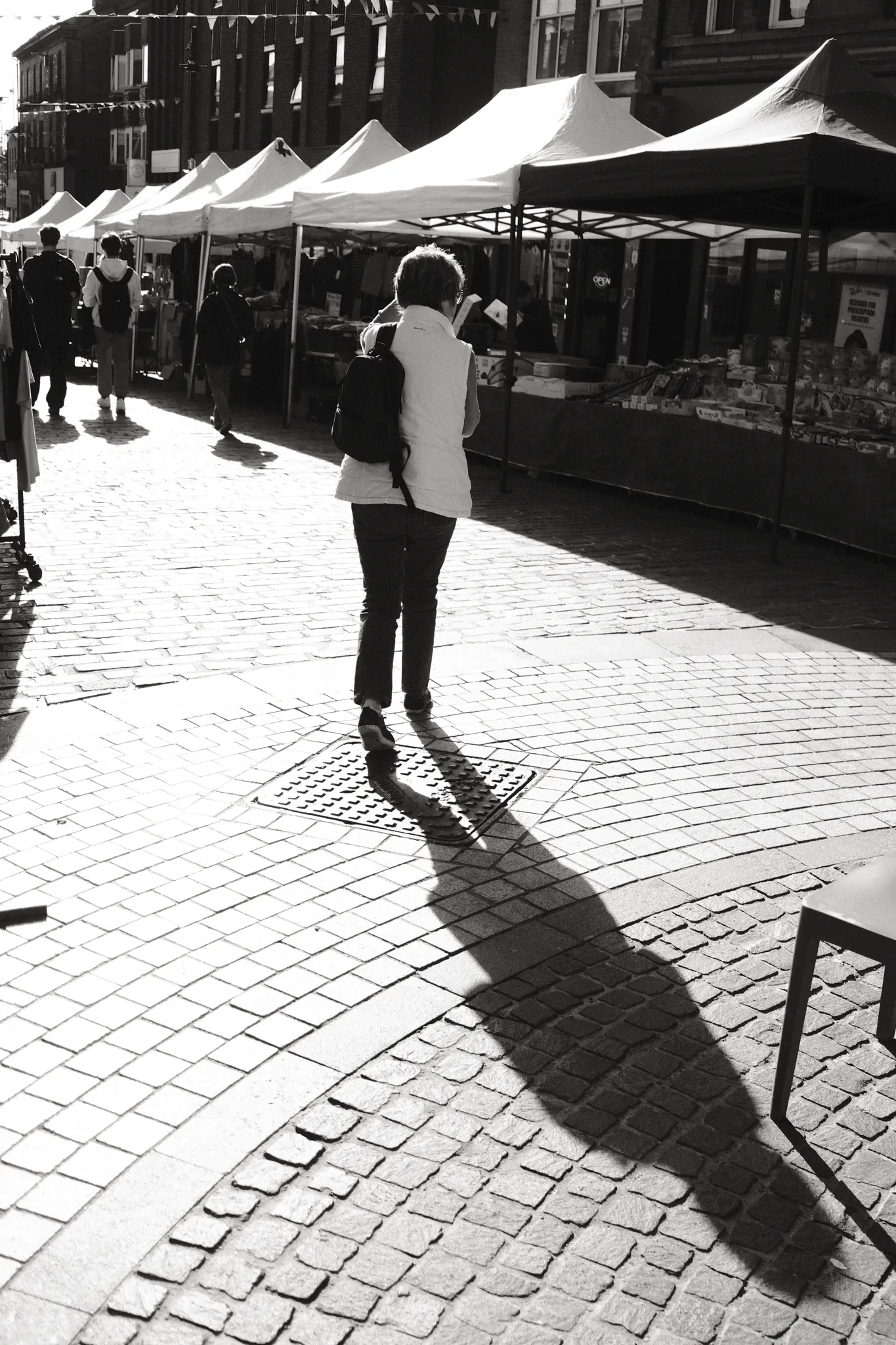 A figure casts a long shadow over the cobbles beside a row of market stalls.