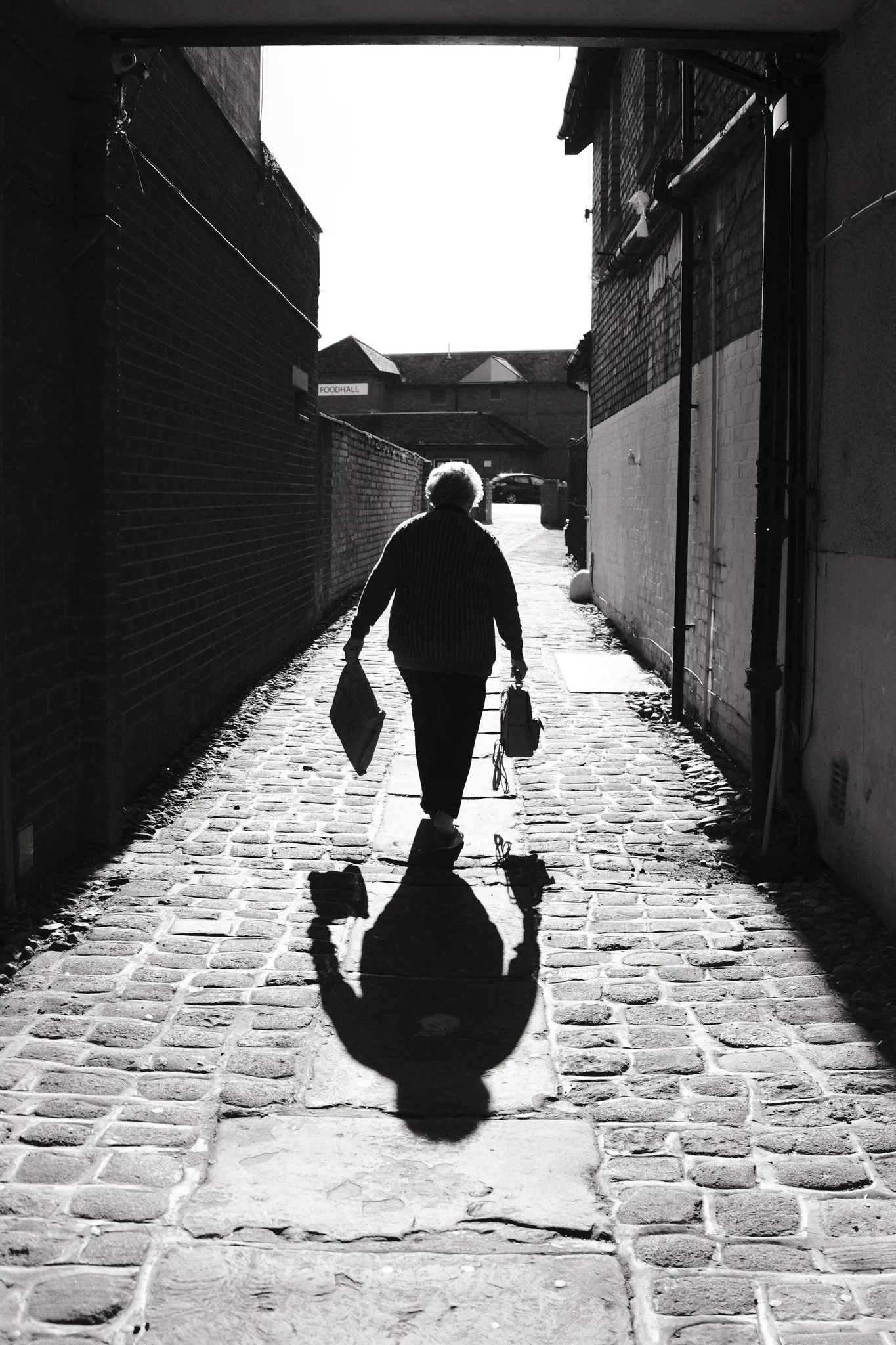 A figure carrying bags walks through a sunlit passage, casting a long shadow over the cobbles.